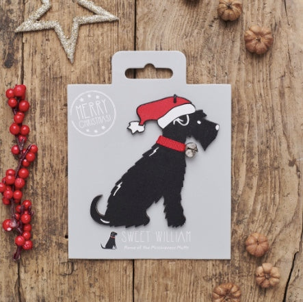 Black dog ornament with a red Santa hat on a white card, surrounded by Christmas decorations on a wooden surface. NZ
